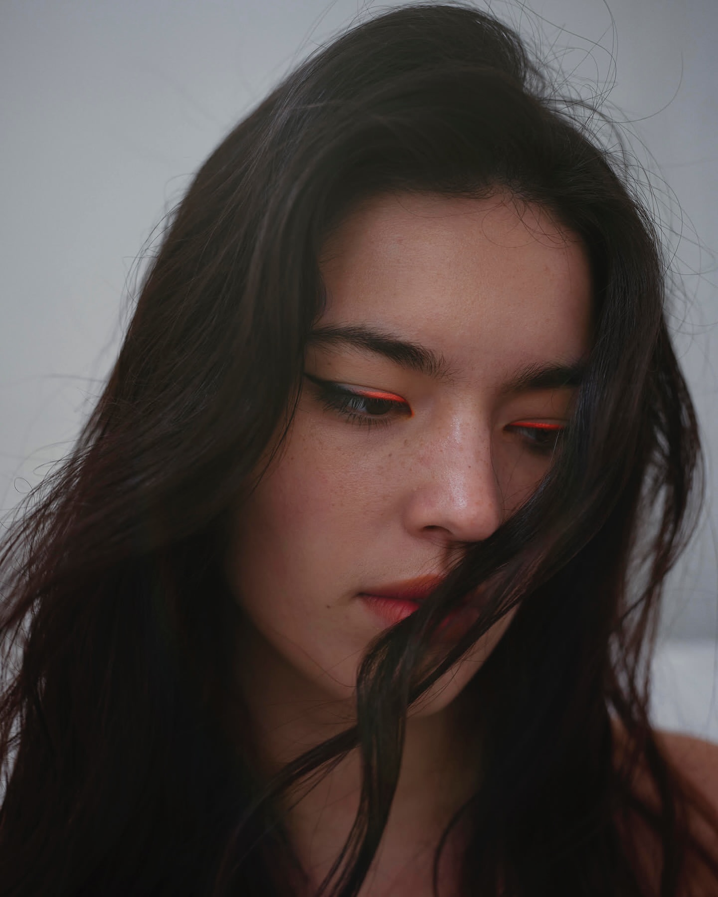 Close-up portrait of a woman with long dark hair and orange eyeliner in a softly lit indoor setting against a plain, bright background.