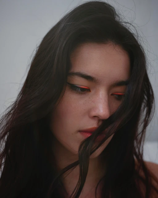Close-up portrait of a woman with long dark hair and orange eyeliner in a softly lit indoor setting against a plain, bright background.