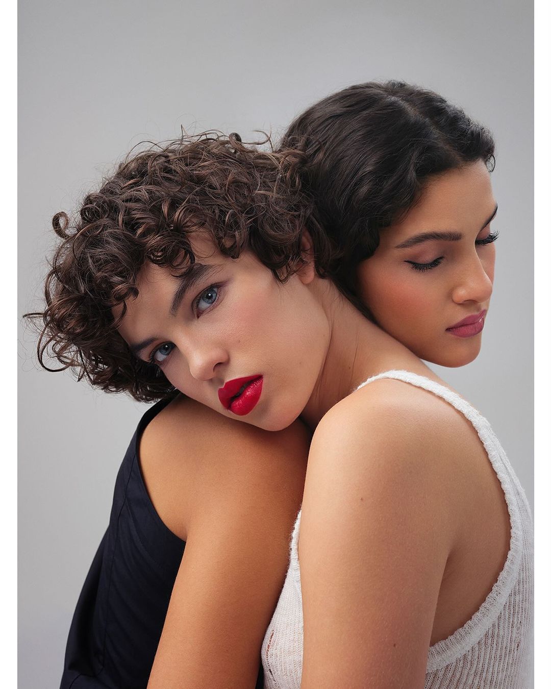 Close-up of two women with elegant makeup, red lipstick, and dark hair. One model wears a knitted tank top, the other a dark sleeveless top.