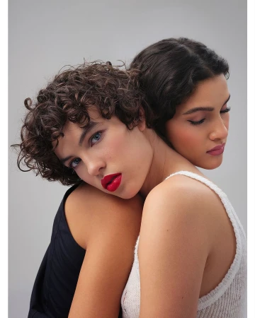 Close-up of two women with elegant makeup, red lipstick, and dark hair. One model wears a knitted tank top, the other a dark sleeveless top.