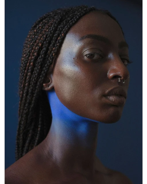 Close-up studio portrait of a Black woman with braids, wearing a nose ring, and with blue-toned lighting against a dark blue background.