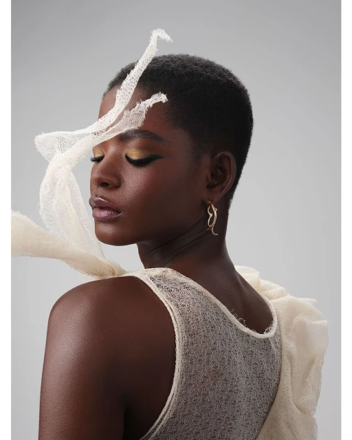 Portrait of a dark-skinned female with short hair wearing sheer white top, decorative headpiece, gold earrings in front of a plain background.