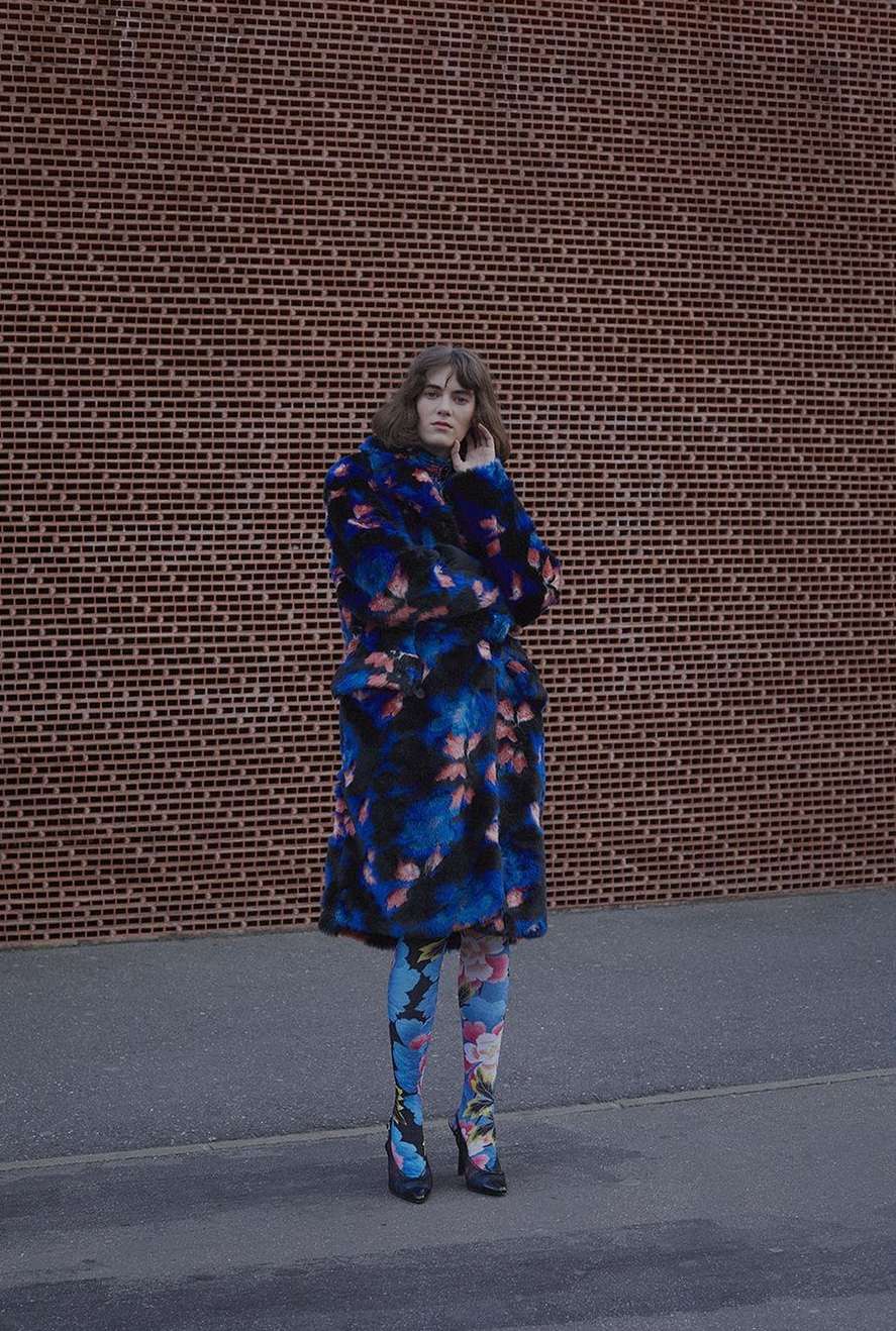 Adult woman with floral coat and leggings poses against a brick wall on paved ground in medium shot, fashion editorial photography.