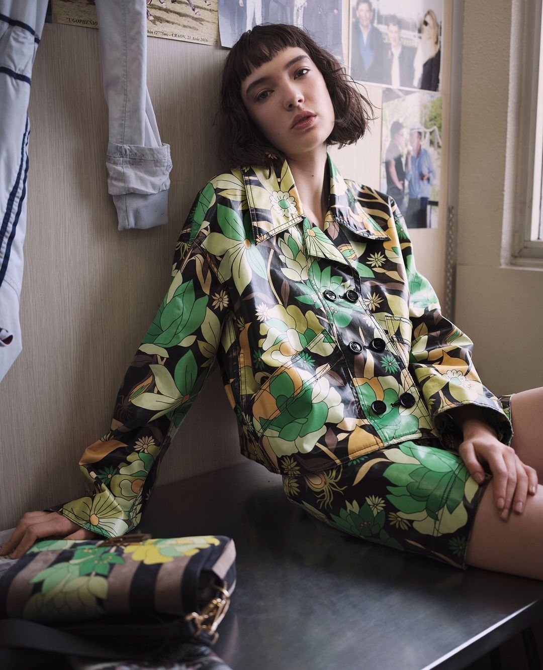 A young woman with bangs in a floral jacket and skirt sits on a surface with a patterned purse indoors with photos in the background.
