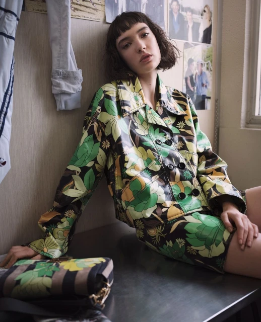 A young woman with bangs in a floral jacket and skirt sits on a surface with a patterned purse indoors with photos in the background.