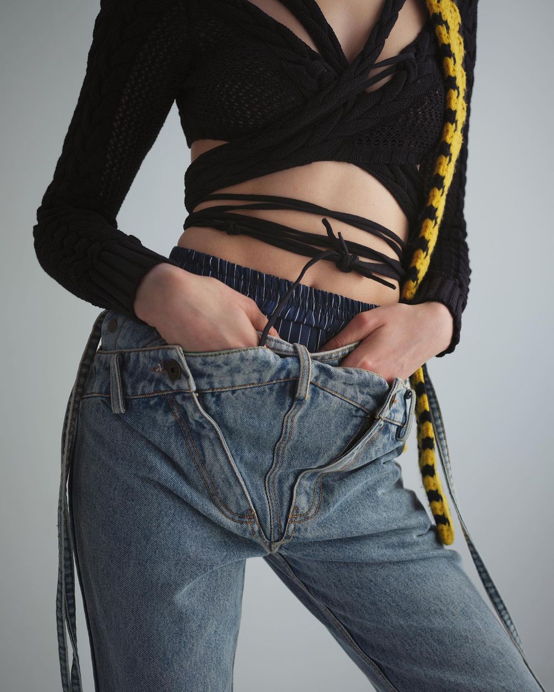 Close-up of a woman wearing a black knit top and light wash jeans with her hands in her pockets, against a neutral backdrop.