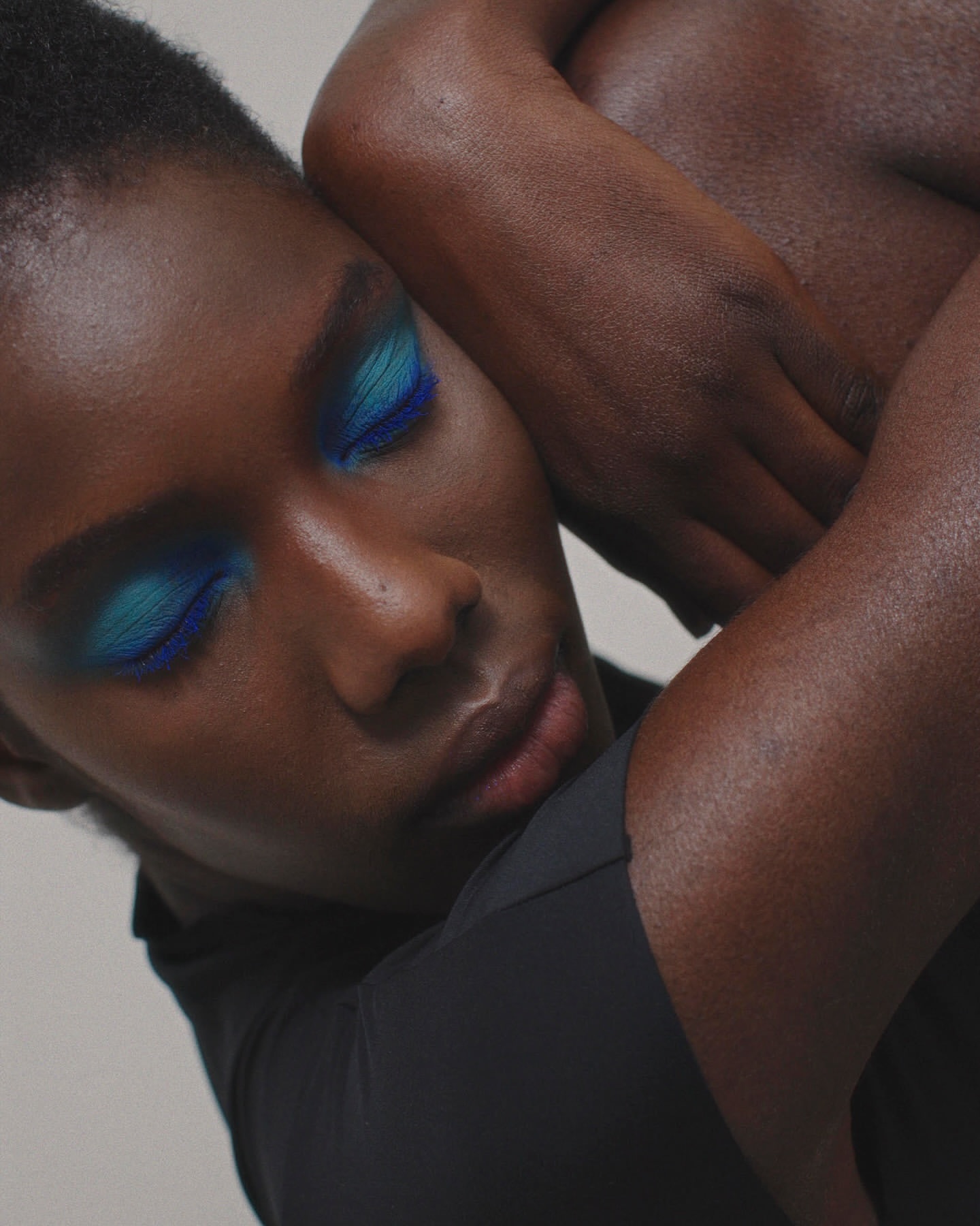 Close-up studio portrait of a dark-skinned person with dramatic blue eyeshadow, leaning against another person, wearing a black top.