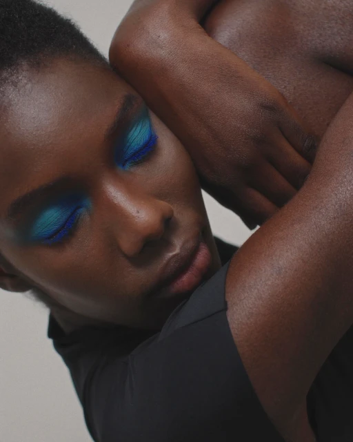 Close-up studio portrait of a dark-skinned person with dramatic blue eyeshadow, leaning against another person, wearing a black top.