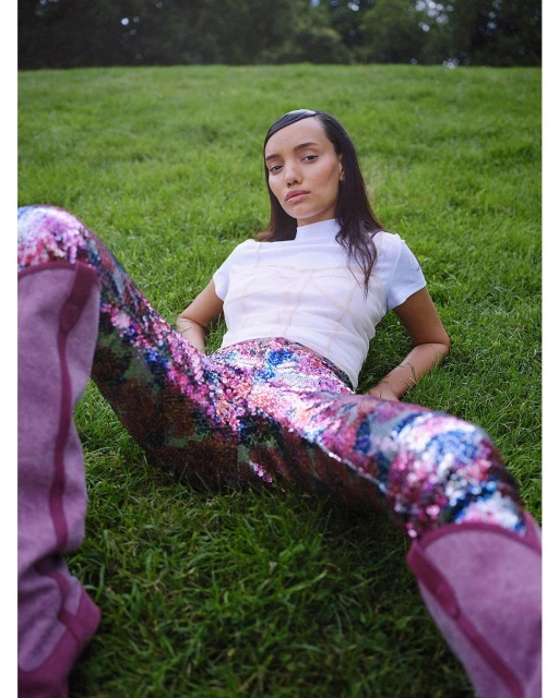Woman in sequined pants lounges on green grass in outdoor setting. She wears a white shirt and boots, gazing directly at the camera.