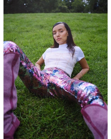 Woman in sequined pants lounges on green grass in outdoor setting. She wears a white shirt and boots, gazing directly at the camera.