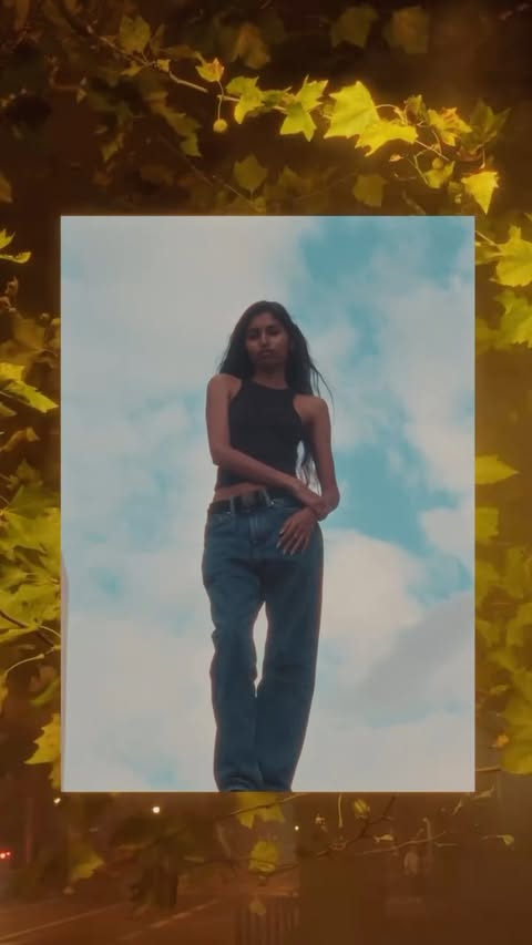 Woman in black tank top and blue jeans standing outdoors against a sky background framed by autumn leaves