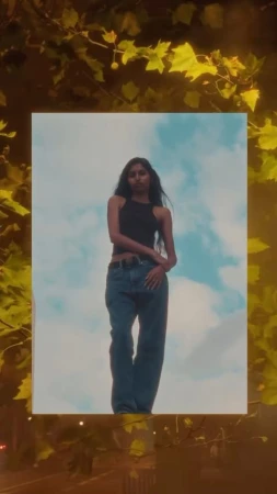 Woman in black tank top and blue jeans standing outdoors against a sky background framed by autumn leaves