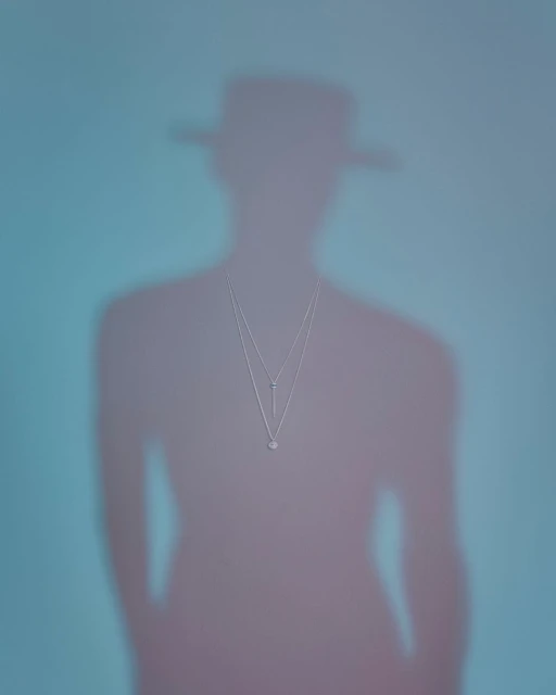 Shadowy figure wearing a hat and a layered necklace on a blue and pink background, jewellery, fashion.