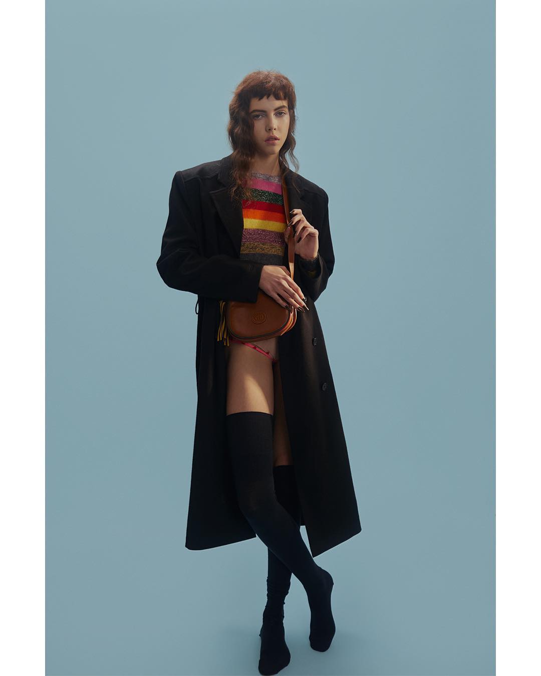 Woman in black coat, colorful sweater, and thigh-high socks poses against a light blue backdrop with a brown shoulder bag.