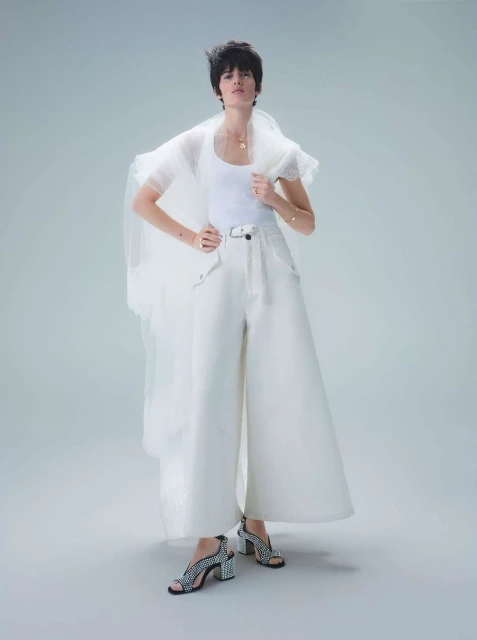 A young woman poses in a white tank top, wide-leg pants, and patterned heels, set against a soft grey backdrop in a fashion shot.
