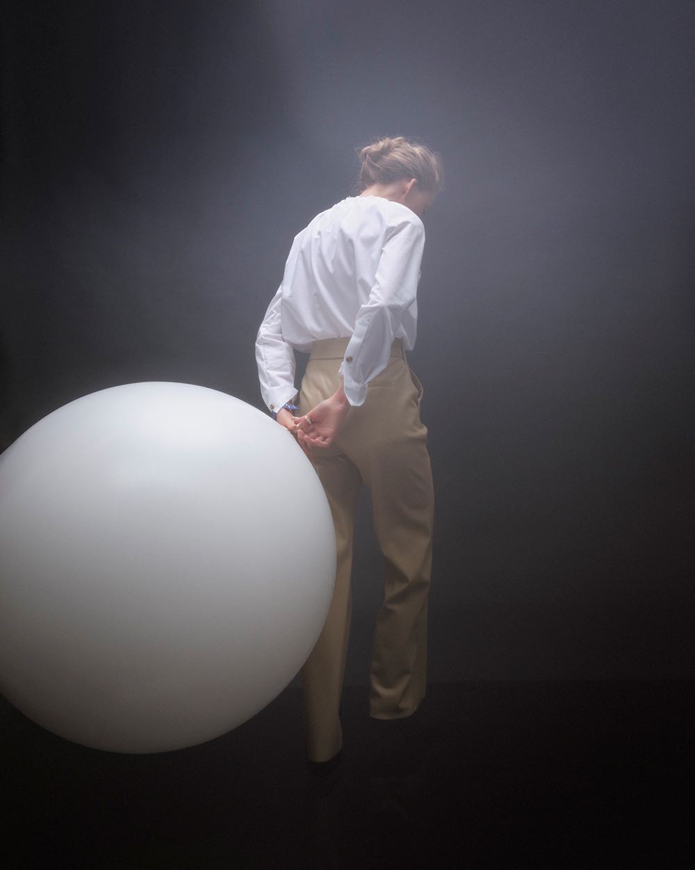 Blonde woman with hair up stands in dark, smoky studio wearing white blouse and tan pants, leaning slightly against a large white sphere.