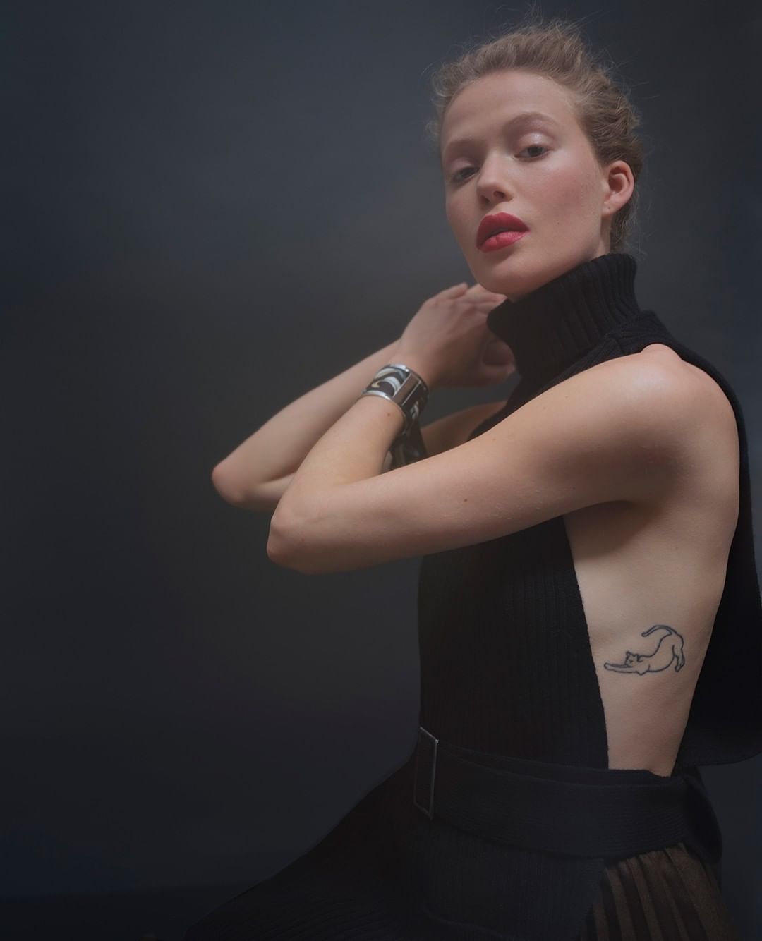 Portrait of a blond adult woman in black high-neck top and skirt, red lipstick, silver bracelet, and a cat tattoo on her side.