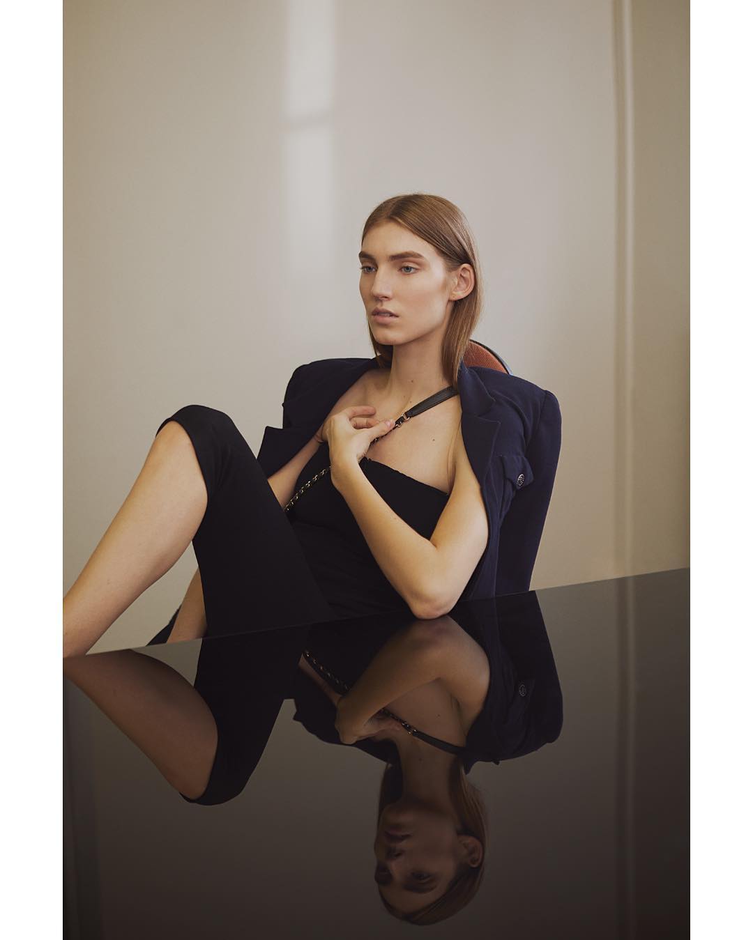Adult woman in a dark tube top and jacket poses in a chair in a studio with a dark, reflective table.