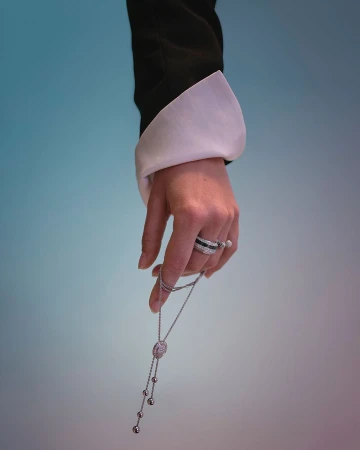 Close-up of a hand wearing diamond rings and holding a pendant necklace against a blue and white gradient background