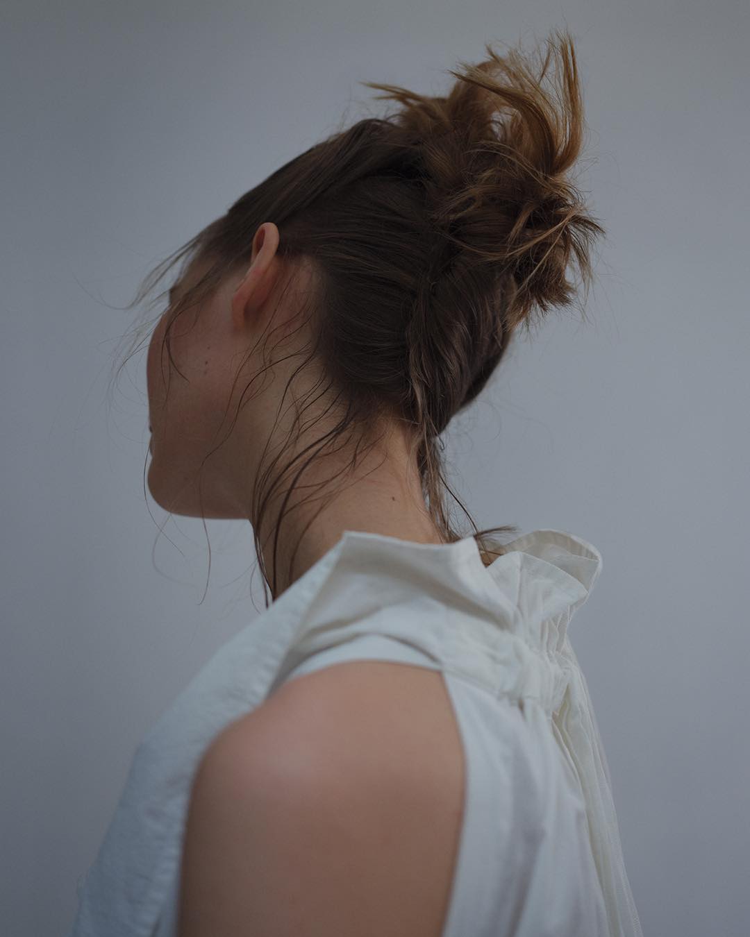 Profile view of a woman with light skin and brown hair styled in a messy bun, wearing a white top against a neutral background.