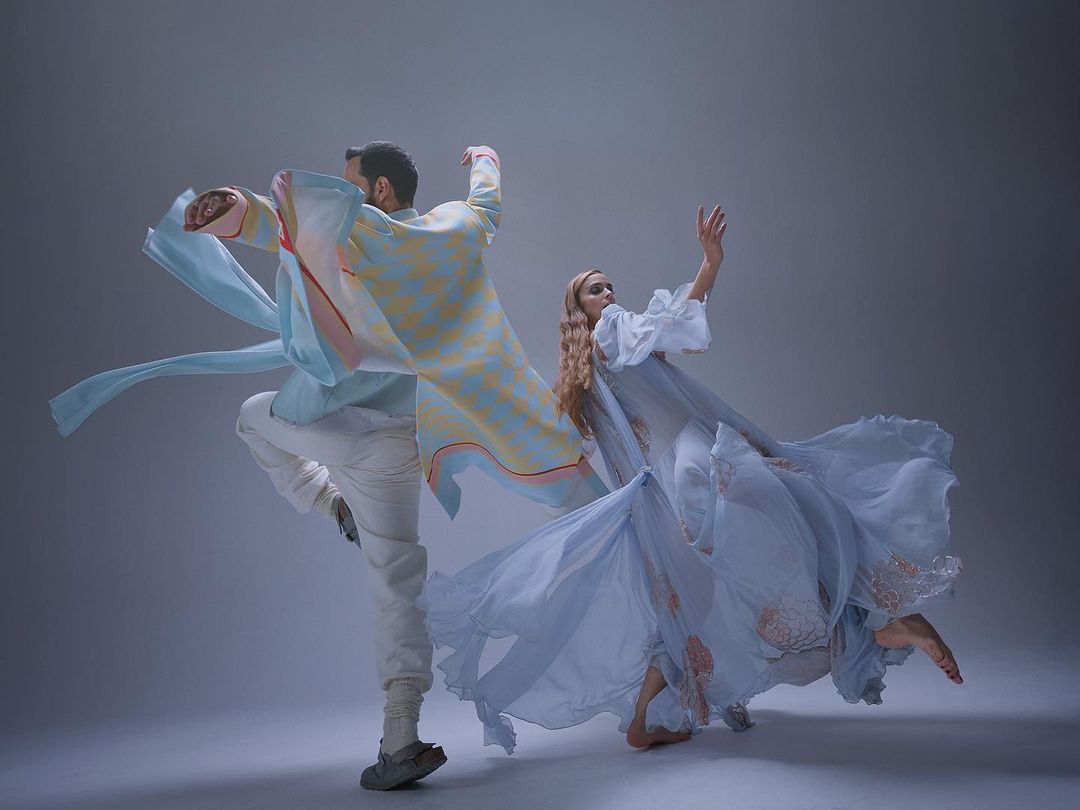 Two dancers in motion, one woman in flowing dress, one man in patterned shirt and white pants, perform against a grey studio backdrop.