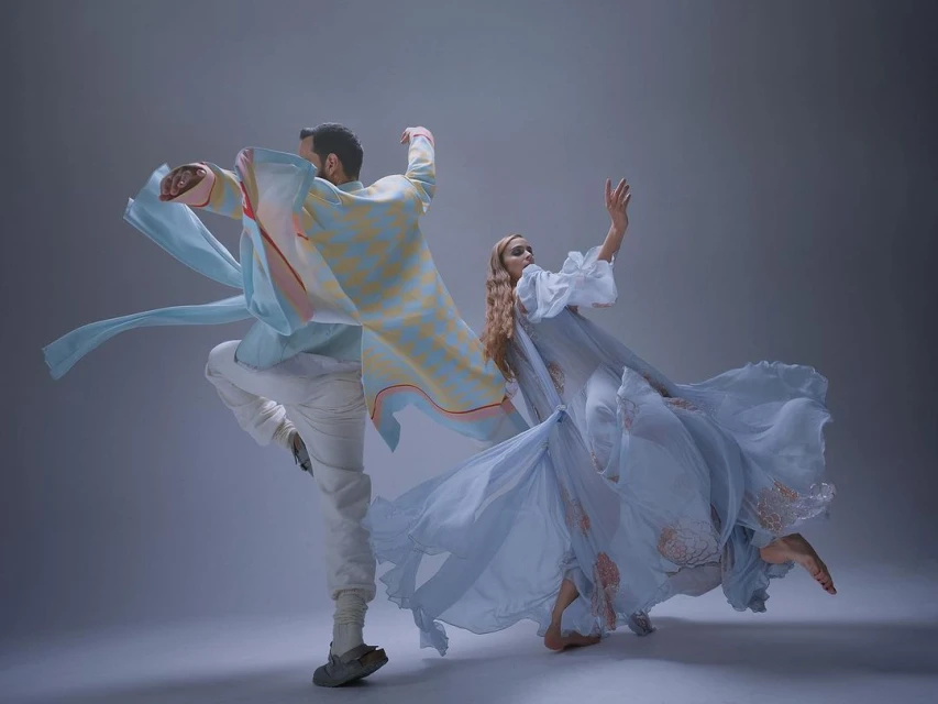 Two dancers in motion, one woman in flowing dress, one man in patterned shirt and white pants, perform against a grey studio backdrop.