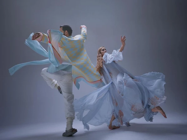 Two dancers in motion, one woman in flowing dress, one man in patterned shirt and white pants, perform against a grey studio backdrop.