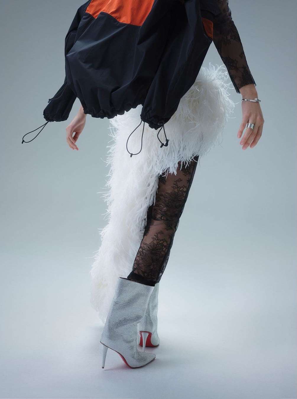 Model in runway fashion wearing black lace tights, white feathered skirt, sparkling boots, and black and orange jacket against white background.