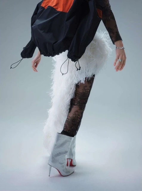 Model in runway fashion wearing black lace tights, white feathered skirt, sparkling boots, and black and orange jacket against white background.