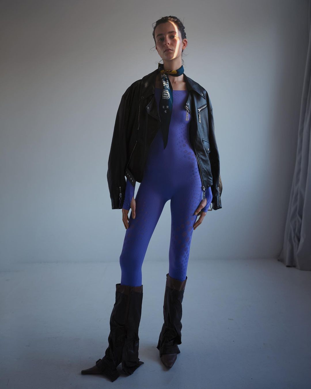 Full-length studio shot of a woman in a purple catsuit, leather jacket, leg warmers, and headscarf posing in front of a neutral backdrop.