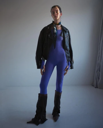 Full-length studio shot of a woman in a purple catsuit, leather jacket, leg warmers, and headscarf posing in front of a neutral backdrop.