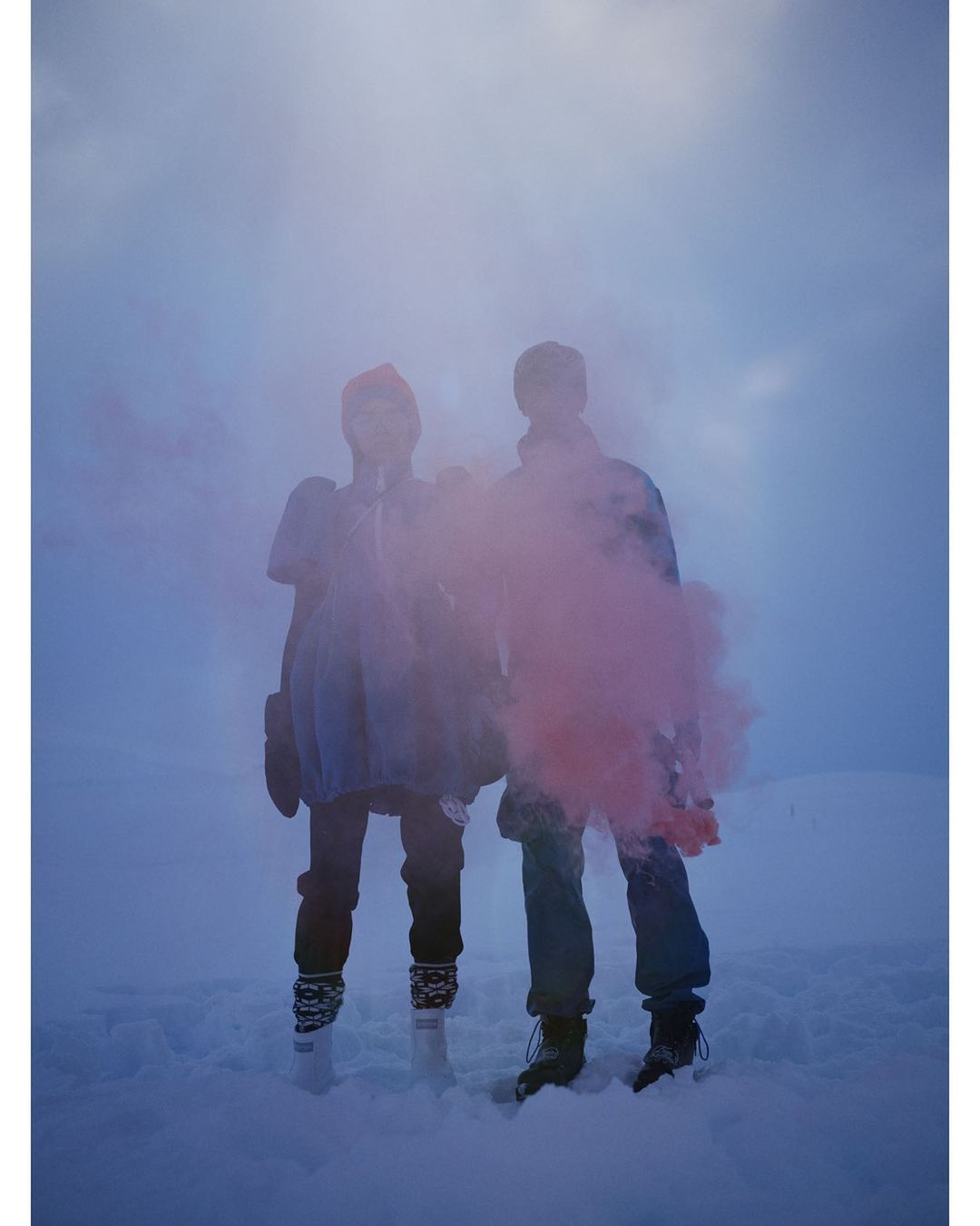 Two people standing in a snowy landscape creating a pink smoke effect against a cloudy sky, wearing winter hats, coats, and boots.