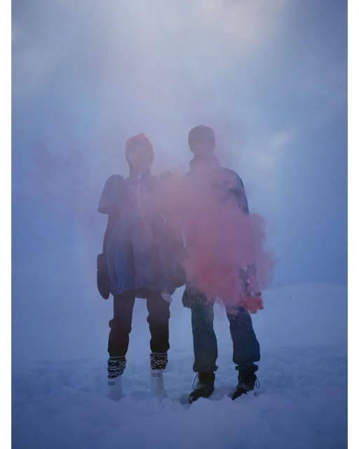 Two people standing in a snowy landscape creating a pink smoke effect against a cloudy sky, wearing winter hats, coats, and boots.