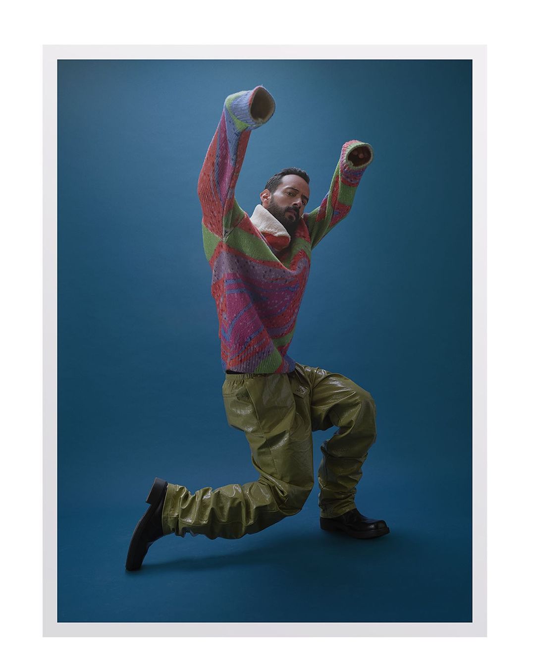 Man wearing a colorful patterned knit sweater and olive green trousers posing in front of a blue background