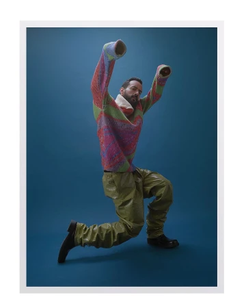 Man wearing a colorful patterned knit sweater and olive green trousers posing in front of a blue background
