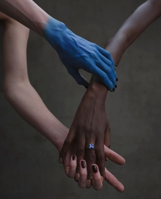 Close-up shot of four human hands intertwined, showcasing painted skin and a ring, set against a blurred grey background.