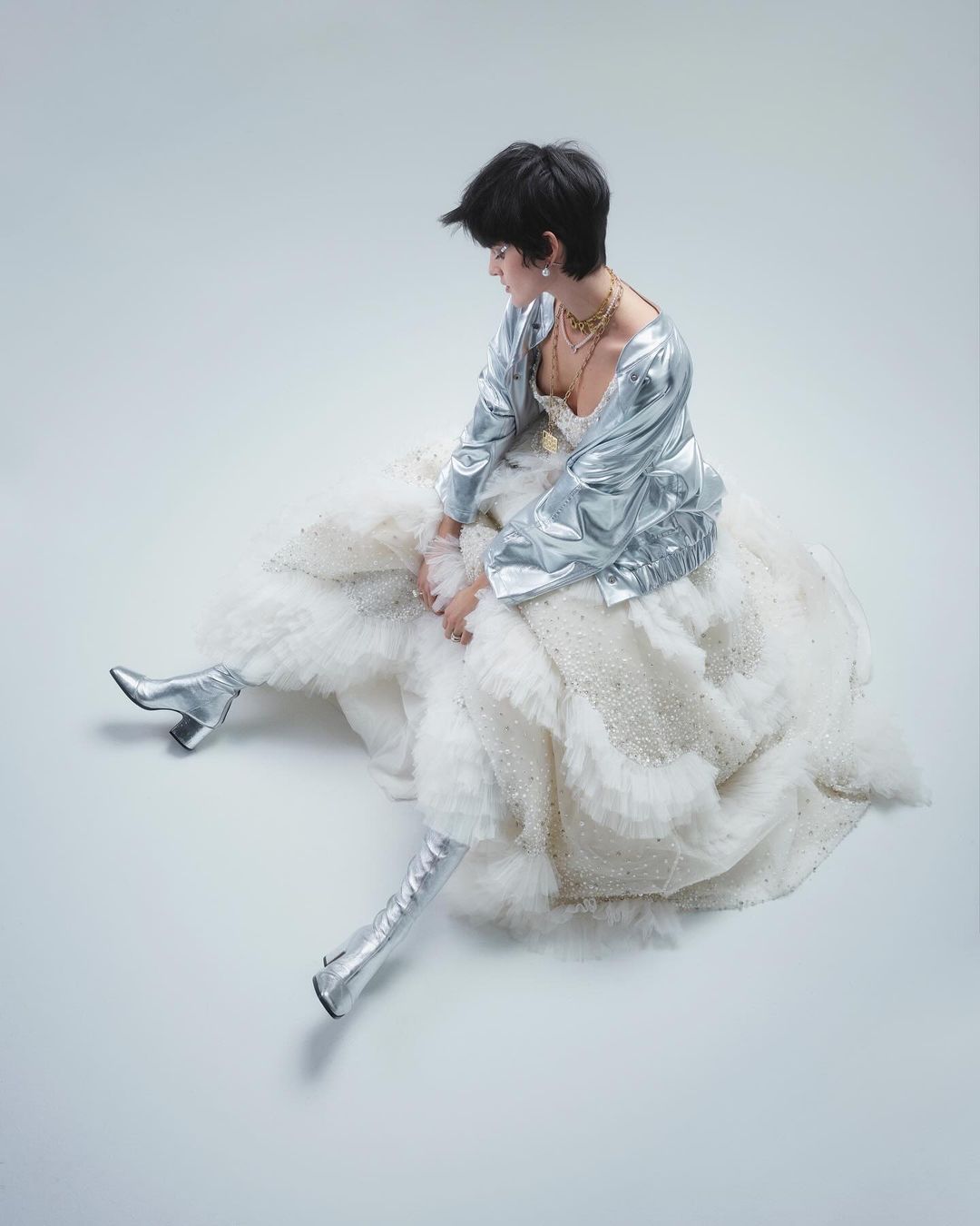 A person sits in a ruffled dress and silver jacket and boots against a seamless background in a fashion or editorial shot.