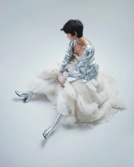 A person sits in a ruffled dress and silver jacket and boots against a seamless background in a fashion or editorial shot.