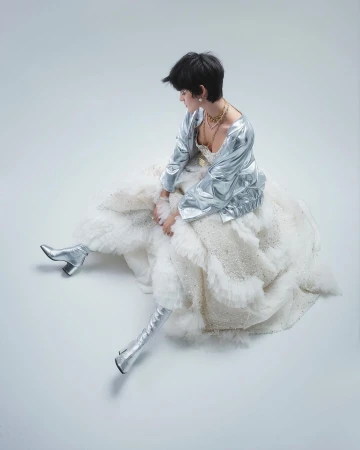 A person sits in a ruffled dress and silver jacket and boots against a seamless background in a fashion or editorial shot.