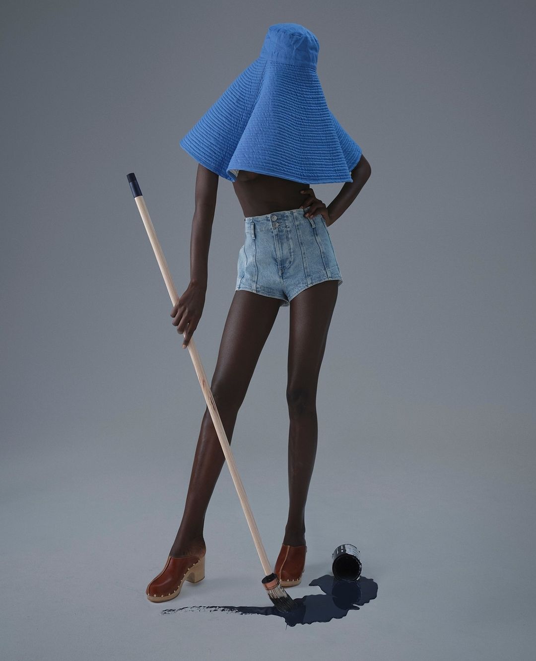Dark-skinned woman in denim shorts and blue hat poses with a paint roller next to a spilled can of dark blue paint indoors on a gray backdrop.