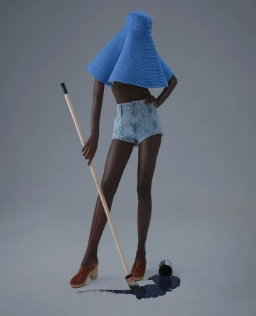 Dark-skinned woman in denim shorts and blue hat poses with a paint roller next to a spilled can of dark blue paint indoors on a gray backdrop.