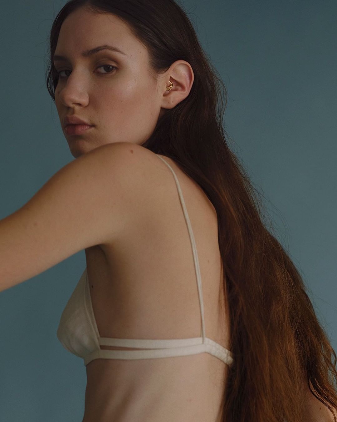 Portrait of a young woman with long brown hair wearing a white top, posing against a blue background in a studio setting.