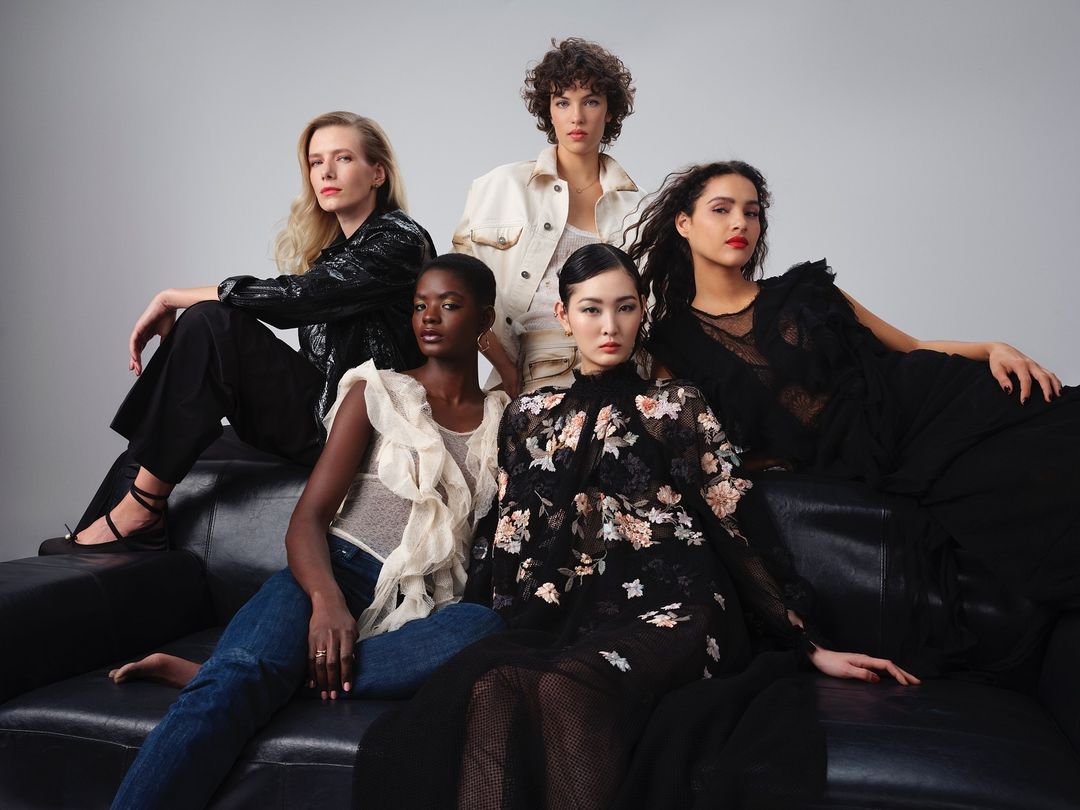 Group portrait of five diverse women seated on a black leather couch, posing in fashionable dresses and stylish tops against a grey background.