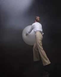Woman in white shirt and beige trousers holding a large white spherical orb under a dramatic overhead spotlight.
