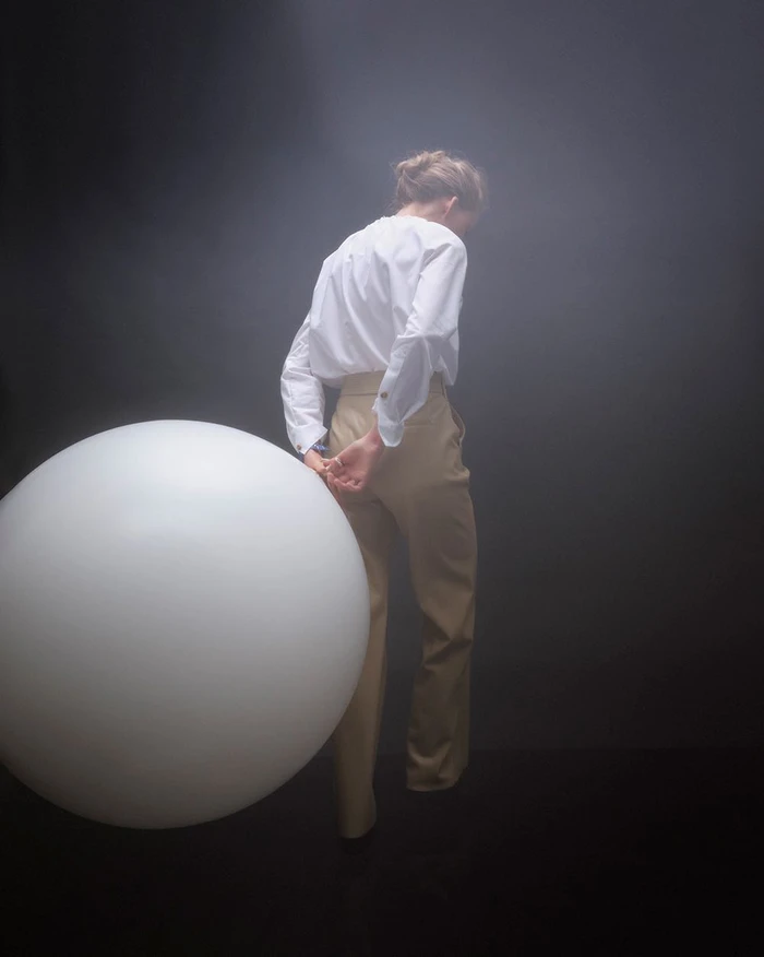 Woman in white shirt and tan trousers stands with a large white sphere in a moody studio by Guillaume Millet