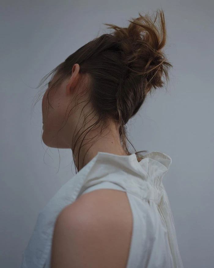 Close-up portrait of a woman with her brown hair in an updo, wearing a white sleeveless top against a grey backdrop.