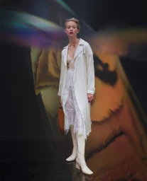 Model in white coat and lace skirt holding a brown bag against a stylized projection background, photographed by Guillaume Millet.