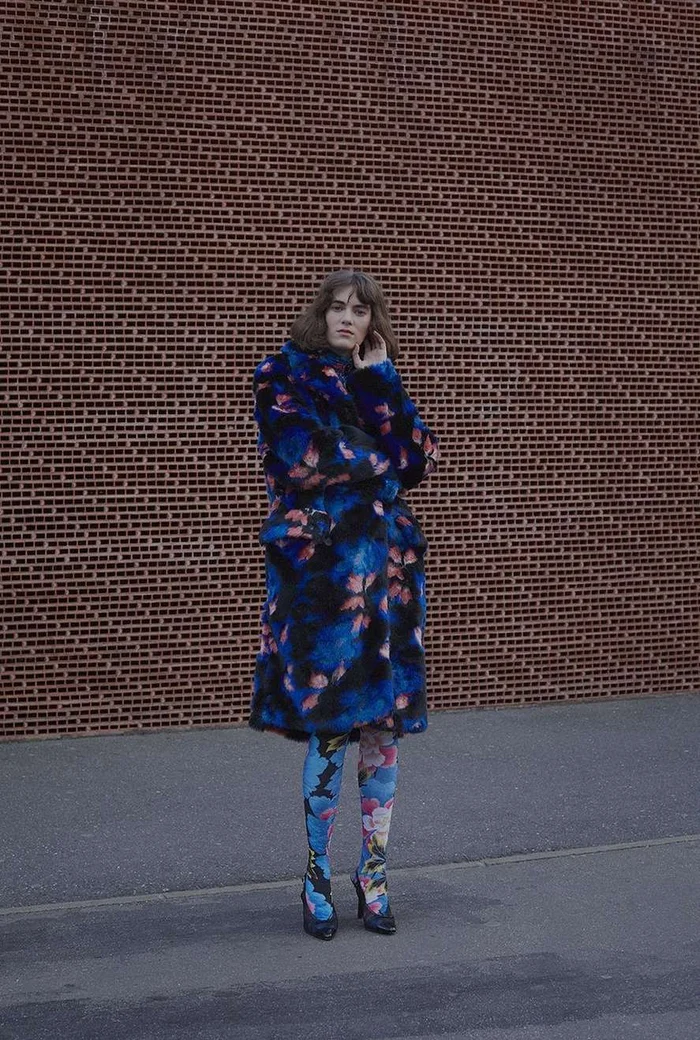 Model in a patterned blue faux fur coat and matching tights standing before a red brick wall