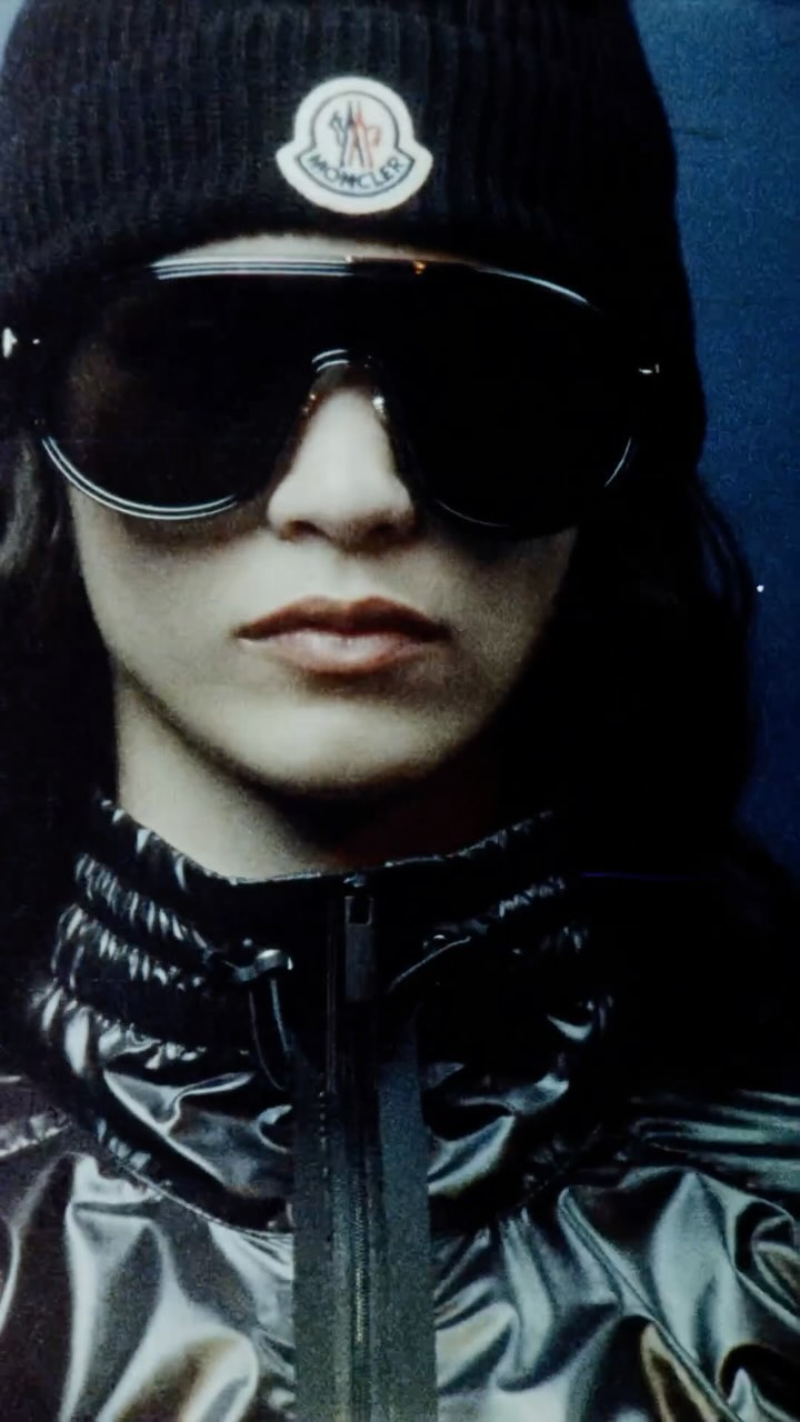 Close up portrait of a model wearing large dark sunglasses, black beanie with logo, and shiny puffer jacket.