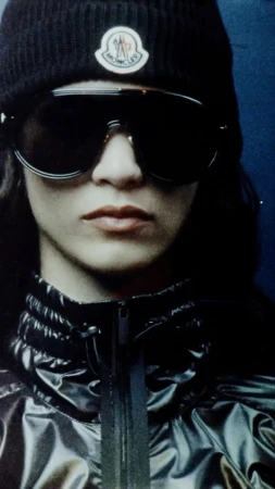 Close up portrait of a model wearing large dark sunglasses, black beanie with logo, and shiny puffer jacket.